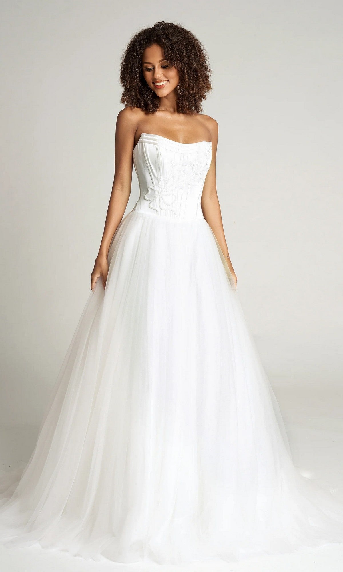 A woman wears the Portia and Scarlett PS26102 strapless ball gown—a white wedding dress with a corset bodice and full tulle skirt—standing against a plain background.